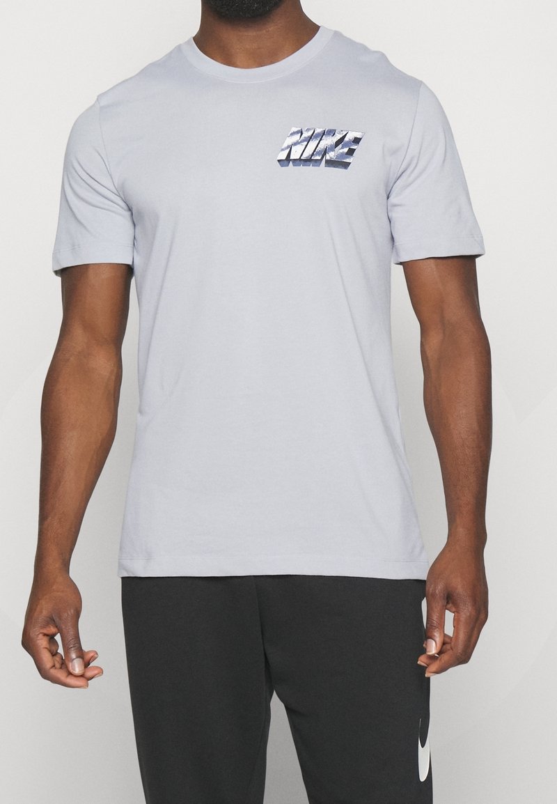 Homme à la peau mate portant un t-shirt Nike gris clair et un pantalon Nike noir, debout devant un fond blanc uni.