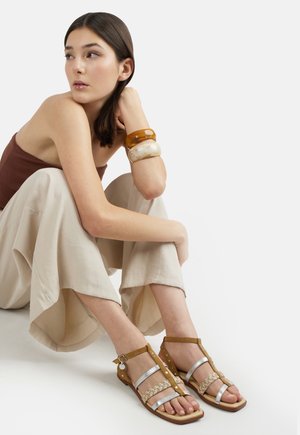 Donna seduta a gambe incrociate che indossa pantaloni beige a gamba larga, canotta marrone, braccialetti ambra e crema e sandali color cuoio con cinturini argentati.