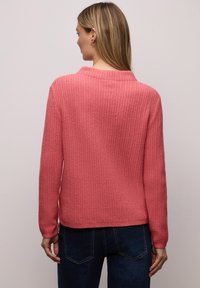Pull côtelé rose à coupe décontractée, manches longues et encolure ronde. Tricot texturé avec une finition douce, porté avec un jean en denim foncé.