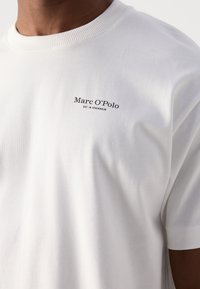Hvit bomulls T-skjorte med rund hals, med en liten svart tekstlogo "Marc O'Polo EST. IN STOCKHOLM" på venstre brystområde.