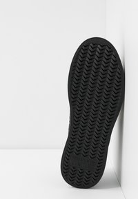 Semelle noire de basket avec motif chevrons, marquée « Versace Jeans Couture » près du talon, positionnée verticalement contre un mur d’angle blanc.