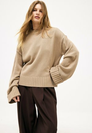 Giovane donna con lunghi capelli castano chiaro che indossa un maglione beige largo e pantaloni scuri a gamba larga, in piedi con una mano sul fianco.