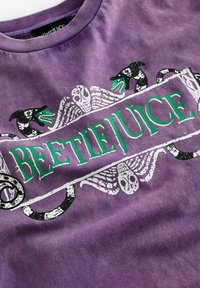 Lila bomullströja med texten "BEETLEJUICE" i grönt, omgiven av svartvita mönster av skallar, vingar och ormar.