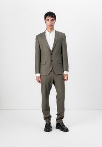 Olive green tailored suit with structured jacket, two buttons, and matching trousers. White button-up shirt and black shoes complete the look.