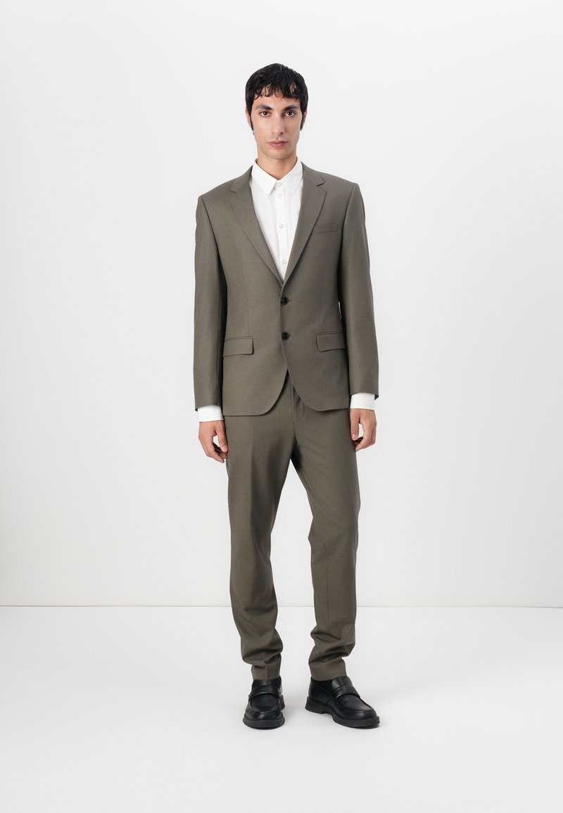 Olive green tailored suit with structured jacket, two buttons, and matching trousers. White button-up shirt and black shoes complete the look.