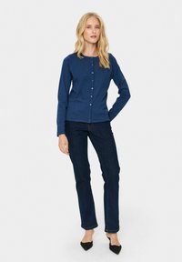 Mornarsko modra jopica z okroglim izrezom in gumbom spredaj; mehak material, dolgi rokavi. Ujemajoča s temno denim hlačami in črnimi špičastimi čevlji.