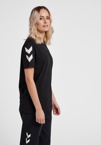 Frau mit schulterlangem blondem Haar, die ein schwarzes T-Shirt und eine Hose mit weißen Chevron-Logos an Ärmeln und Hosenbein trägt, seitlich stehend.