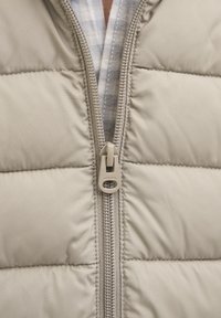 Chaqueta acolchada beige con el zipper parcialmente abierto, revelando una tela a cuadros en tonos azul claro y blanco debajo.