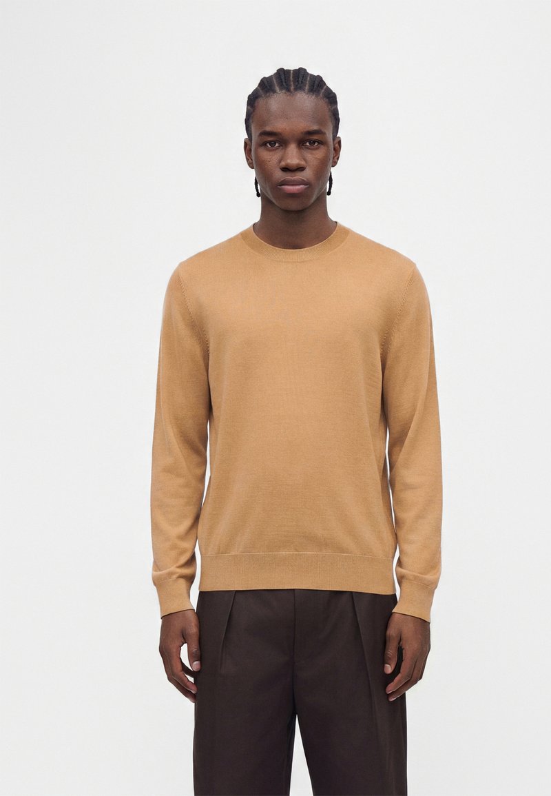 Homme aux cheveux tressés, portant un pull beige à manches longues et un pantalon marron foncé, debout devant un fond clair et uni.