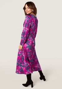 Robe à manches longues et mi-longue de couleur rose vif avec des motifs floraux bleus et violets, fabriquée en tissu léger, associée à des bottes à talons noires.