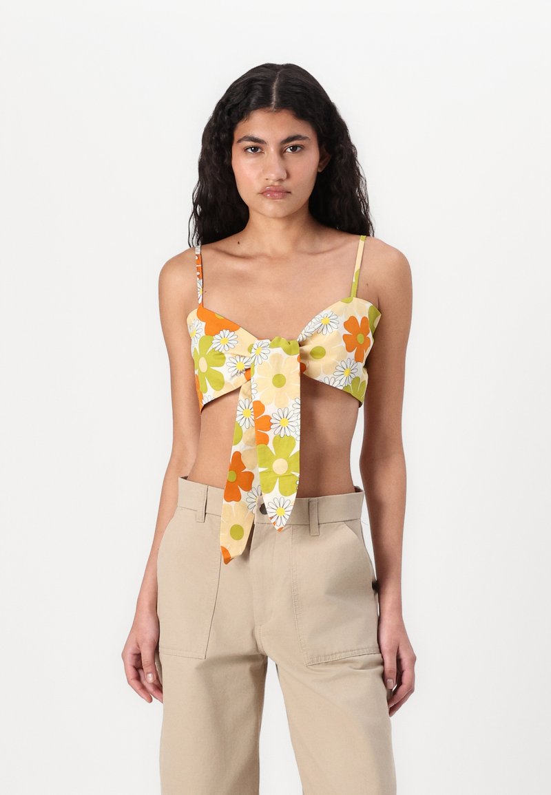Top corto floreale giallo con fiori arancioni, verdi e bianchi. Caratterizzato da spalline sottili e un dettaglio annodato sul davanti, abbinato a pantaloni beige.