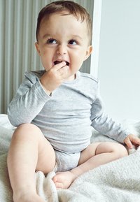 Grå langærmet skjorte med rund hals, lysegrå shorts, siddende på et tekstureret tæppe, holder en lille snack, smilende.