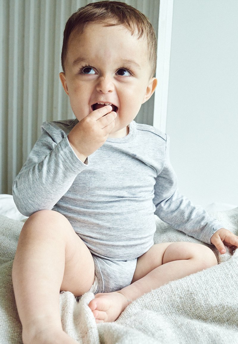 Grå langærmet skjorte med rund hals, lysegrå shorts, siddende på et tekstureret tæppe, holder en lille snack, smilende.