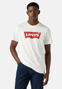 Ung mand iført en hvid Levi's t-shirt med rødt logo, stående med den ene hånd i lommen og en neutral udtryk.