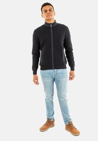 Suéter de punto azul marino con cremallera y cuello alto, que presenta bolsillos laterales grises y dobladillo acanalado, combinado con vaqueros de color azul claro y botas marrones.