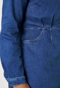 Camisa de denim en azul oscuro con un diseño ajustado y elegante, costuras visibles, un bolsillo lateral y mangas remangadas.