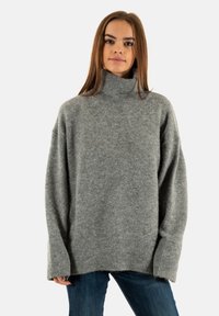 Pull en maille gris à col roulé avec une coupe décontractée, des poignets et un ourlet côtelés. Texture douce et design minimaliste. Porté avec un jean bleu.