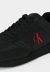 Baskets basses noires avec lacets noirs, logo rouge « cK » sur le côté et semelle noire plate.