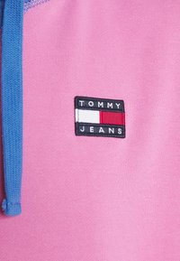 Tessuto rosa con cordino blu e una patch rettangolare nera con il testo "TOMMY" e "JEANS" affiancato da strisce rosse, bianche e blu.