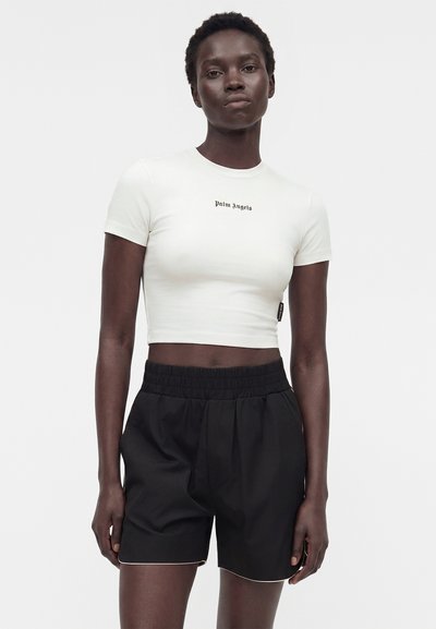 Γυναίκα που στέκεται με το πρόσωπο προς τα εμπρός, φοράει ένα λευκό κοντό μπλουζάκι με το κείμενο "Palm Angels" και μαύρο σορτς με ελαστική μέση σε λευκό φόντο.