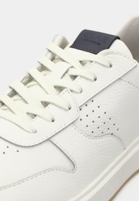 White leather sneaker with white laces, perforated side detail, and dark label on tongue against a plain white background.