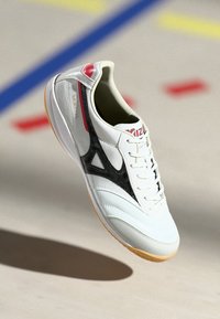 Witte sportschoen met zwarte accenten, gemaakt van leer en synthetische materialen, vetersluiting, rubberen zool, met een rood logo en unieke patronen.