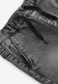 Graue Denim-Hosen mit elastischem Bund, schwarzen Kordeln und einem kleinen schwarzen Logoschild. Die Textur wirkt leicht abgenutzt und weich.