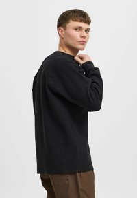 Top negru cu mâneci lungi dintr-un material texturat, cu guler rotund și umeri coborâți, asociat cu pantaloni maronii. Design simplu, fără logo-uri.