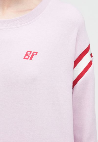 Φούτερ απαλό ροζ με κόκκινο λογότυπο "BP" στο στήθος και δύο κόκκινες και λευκές ρίγες στο δεξί μανίκι.
