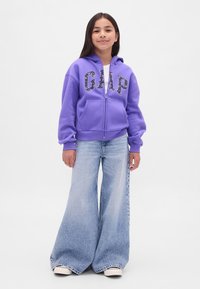 Camisola com capuz roxa com fecho de correr e logótipo "GAP", combinada com jeans de perna larga azul-claro. A camisola tem bolsos frontais e punhos canelados. Ténis brancos.