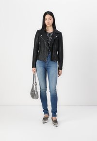 Chaqueta de biker de cuero negro sobre una blusa con estampado en blanco y negro, combinada con jeans ajustados azul claro y mocasines plateados. Sosteniendo un bolso plateado.