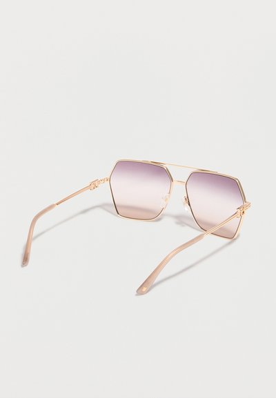 Γυαλιά ηλίου με εξαγωνικό μεταλλικό χρυσό πλαίσιο, φακούς με βαθμιαία μοβ χρώμα και λεπτούς βραχίονες, τοποθετημένα σε λευκή επιφάνεια.