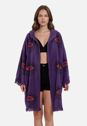 Jonge vrouw die een losse paarse jas met rode en blauwe oogpatronen draagt, een zwart crop topje en zwarte korte broek, staand voor een effen achtergrond.