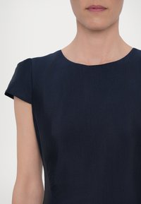 Tummansininen mekko, jossa on sileä pinta, hihatettomat hihat ja pyöreä pääntie. Mekossa on istuva yläosa ja hienovarainen sivuruuska.