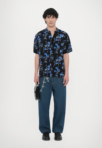 Svart kortärmad skjorta med blå blommönster, kombinerad med lösa blåjeans med dekorationer, och svarta skor, bärandes en liten väska.