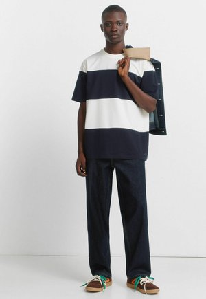 Jonge man die een marineblauw-wit gestreept t-shirt draagt, donkere spijkerbroek, kleurrijke sneakers, een spijkerjack over zijn schouder houdt, staand tegen een witte muur.