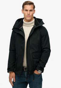 Schwarze wasserdichte Jacke mit Kapuze, zwei Reißverschluss-Taschen, verstellbaren Bündchen, kombiniert mit einem beigen Strickpullover und blauen Jeans.