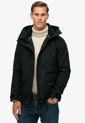 Zwarte waterdichte jas met een capuchon, twee ritszakken, verstelbare manchetten, gecombineerd met een beige gebreide trui en blauwe jeans.