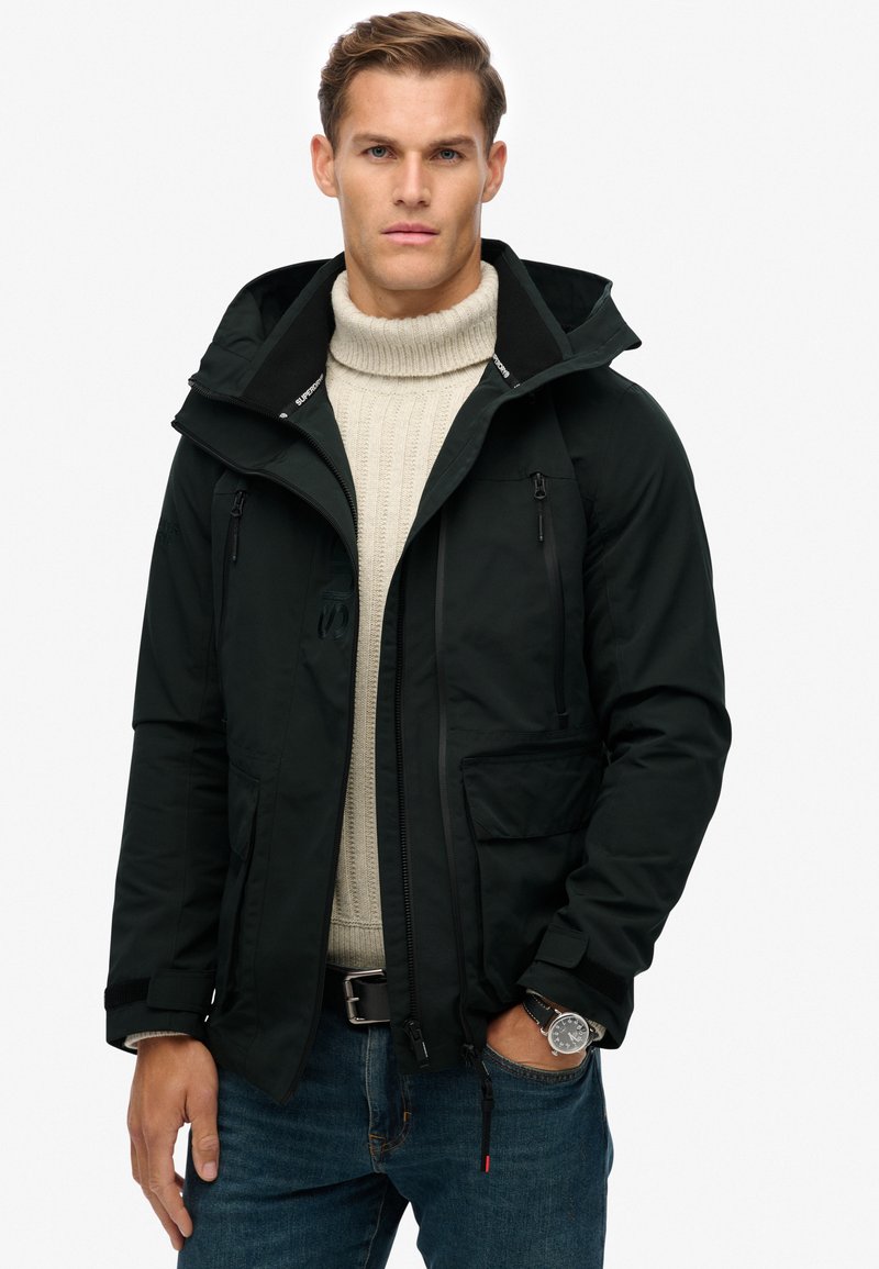 Schwarze wasserdichte Jacke mit Kapuze, zwei Reißverschluss-Taschen, verstellbaren Bündchen, kombiniert mit einem beigen Strickpullover und blauen Jeans.