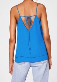 Top en camisole bleu avec des bretelles spaghetti réglables et un dos en V profond, présentant un détail à nouer et un ourlet superposé sur un jean en denim clair.