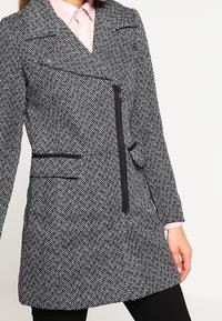 Abrigo de patrón de espiga en blanco y negro, con un diseño cruzado, cierre con cremallera y bolsillos laterales, con un ajuste entallado.