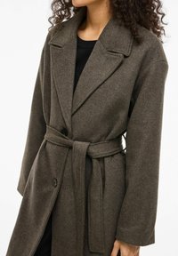 Ruskea villasekoitekate, jossa on vyötärölle solmittava vyö, leveät kaulukset ja suuret napit; siinä on rento istuvuus ja pitkät hihat.