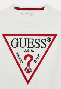 Biele tričko z organickej bavlny s výrazným červeným trojuholníkovým logom, na ktorom je čiernym vyšívaním napísané "GUESS," "WASHED" a "JEANS" a dekoratívny otáznik.