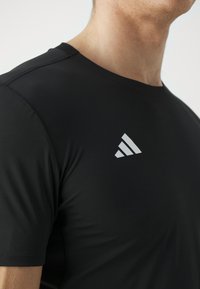 Camiseta atlética negra de tela suave, con mangas cortas y un logo de tres franjas blancas en el área izquierda del pecho.