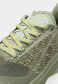 Close-up van een lichtgroene hardloopschoen met veters met gele strepen en een geometrisch overlay-ontwerp op het zijpaneel.