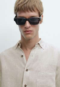 Schwarze und schildpattfarbene Sonnenbrille mit quadratischen Rahmen. Beiges Leinenhemd mit kurzen Ärmeln, Knopfkragen und einer Brusttasche.