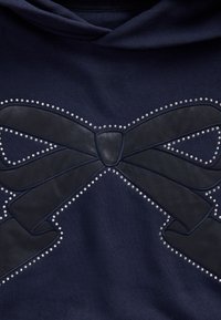 Granatowa bluza z dużą skórzaną kokardą ozdobioną srebrnymi ćwiekami, które kontrastują z tkaniną.