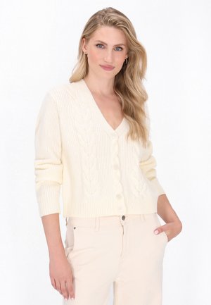 Crème kleurige cardigan met V-hals, kabelbreistekstuur, lange mouwen en knopen. Gedragen over lichtbeige broeken.