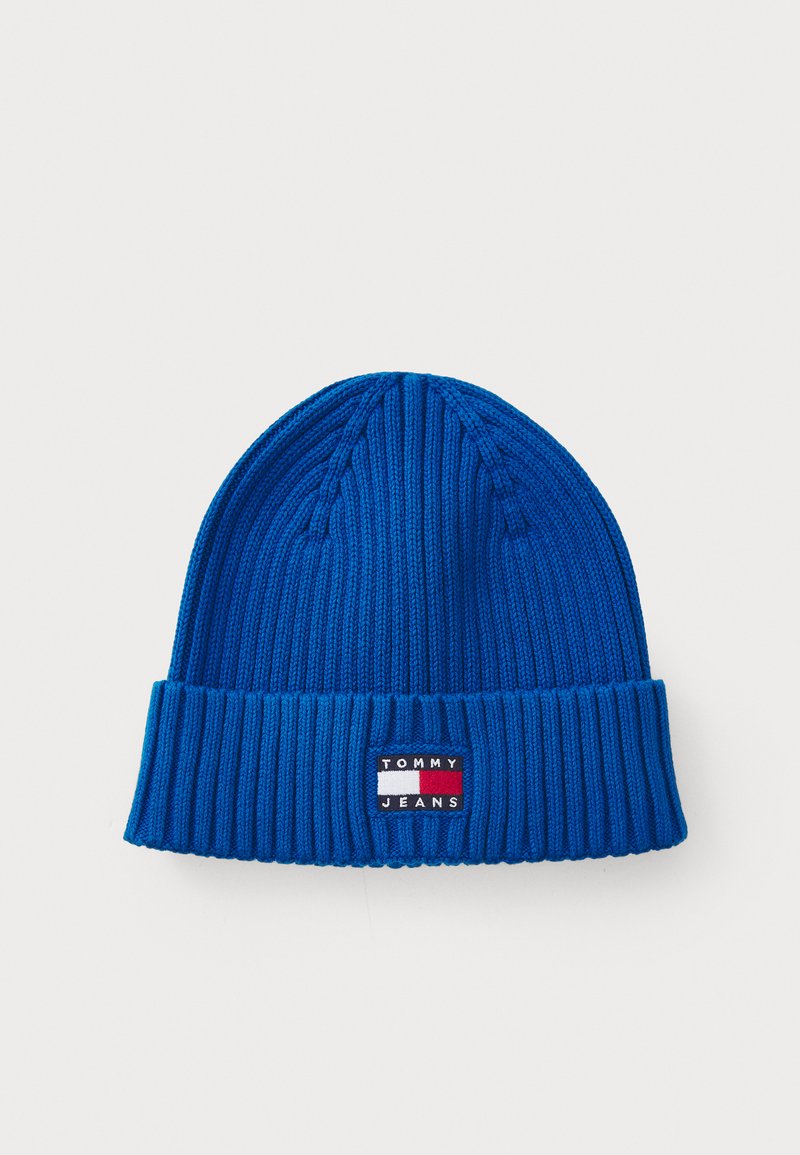 Blå ribbad stickad mössa med en vikbar kant och en rektangulär logotyp-patch med röda, vita och blå ränder.