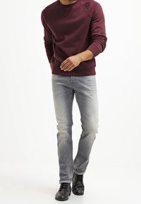 Uomo che cammina indossando un felpa bordeaux, jeans grigio chiaro e scarpe di pelle nere su uno sfondo bianco.